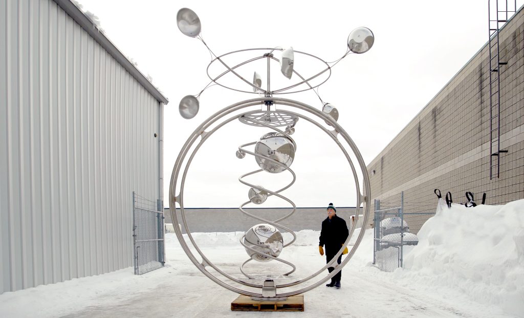 A man standing looking a a circular steel kinetic sculpture outside a warehouse. 