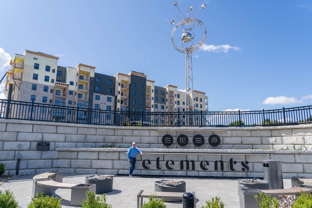 A man stands in front of a modern apartment complex named “Elements,” featuring symbols of earth, water, fire, air, and spirit. A futuristic metallic sculpture stands tall behind him.